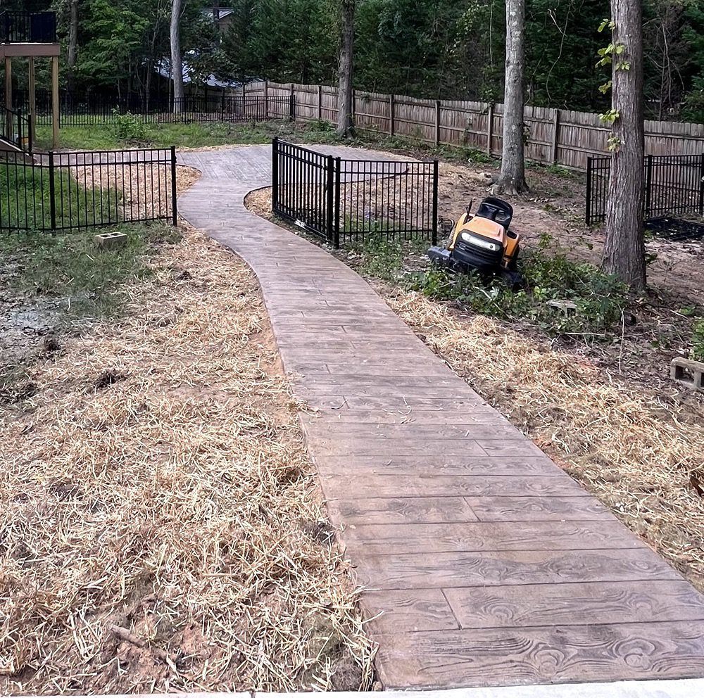 A concrete walkway with a wood-plank texture curves through a yard featuring black metal fencing and wood mulch.
