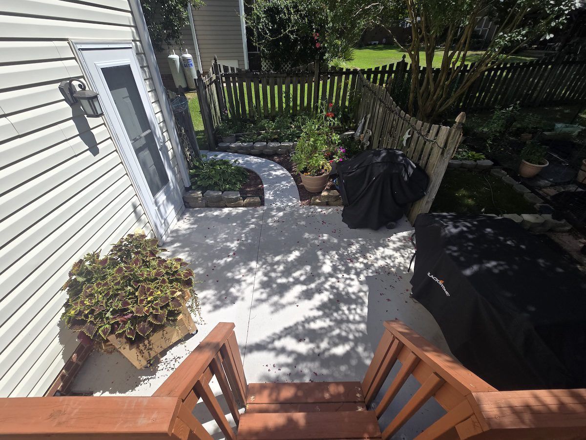 A patio view from wooden stairs, showing a concrete area with potted plants, a covered grill, and a fenced yard.