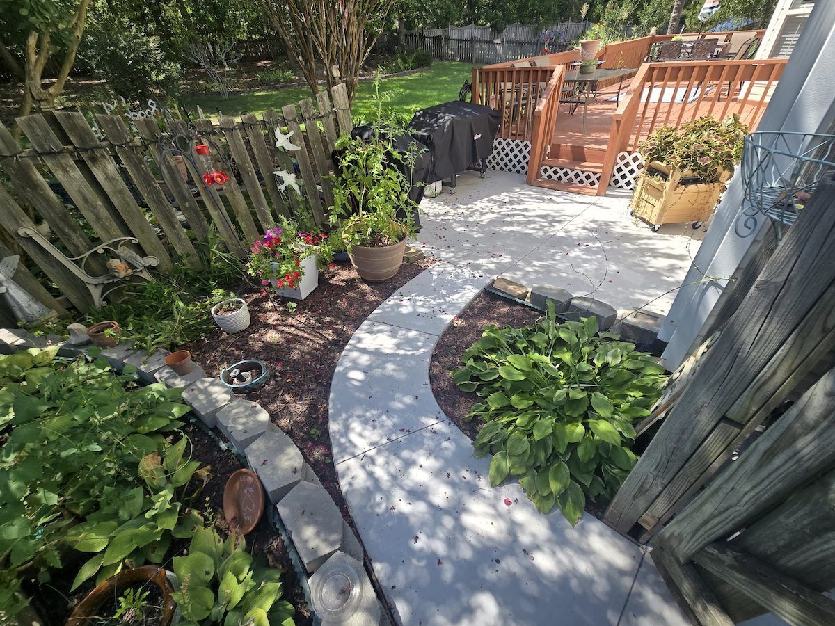 A high-angle view of a backyard featuring a curved gray concrete path, garden beds, potted plants, and a wooden deck.