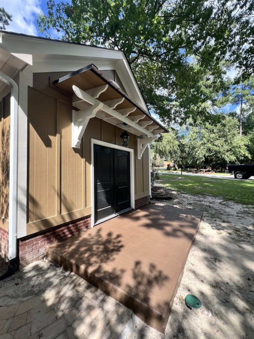A brown building with a black double door, a decorative white awning, and a small concrete landing in a wooded setting.