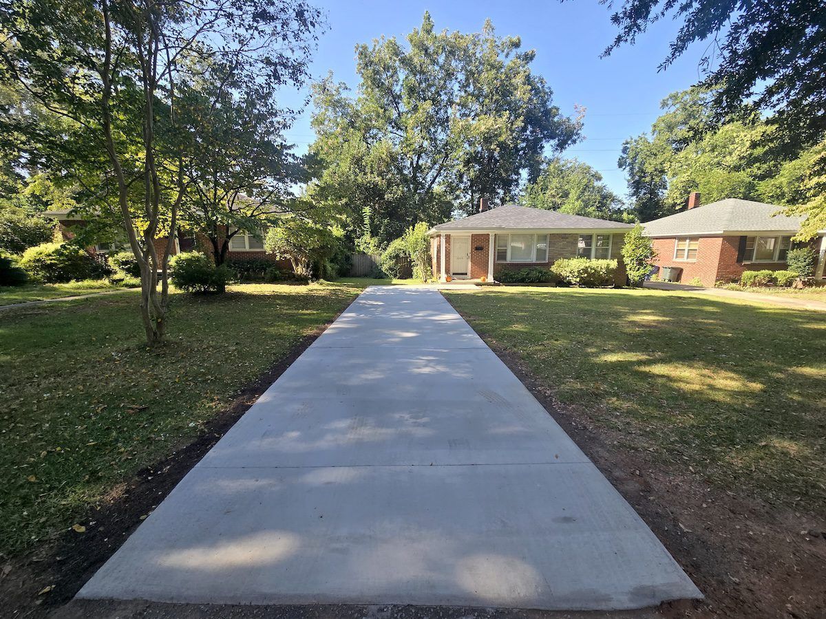 A new concrete driveway stretches from the foreground toward two brick houses surrounded by trees on a sunny day.