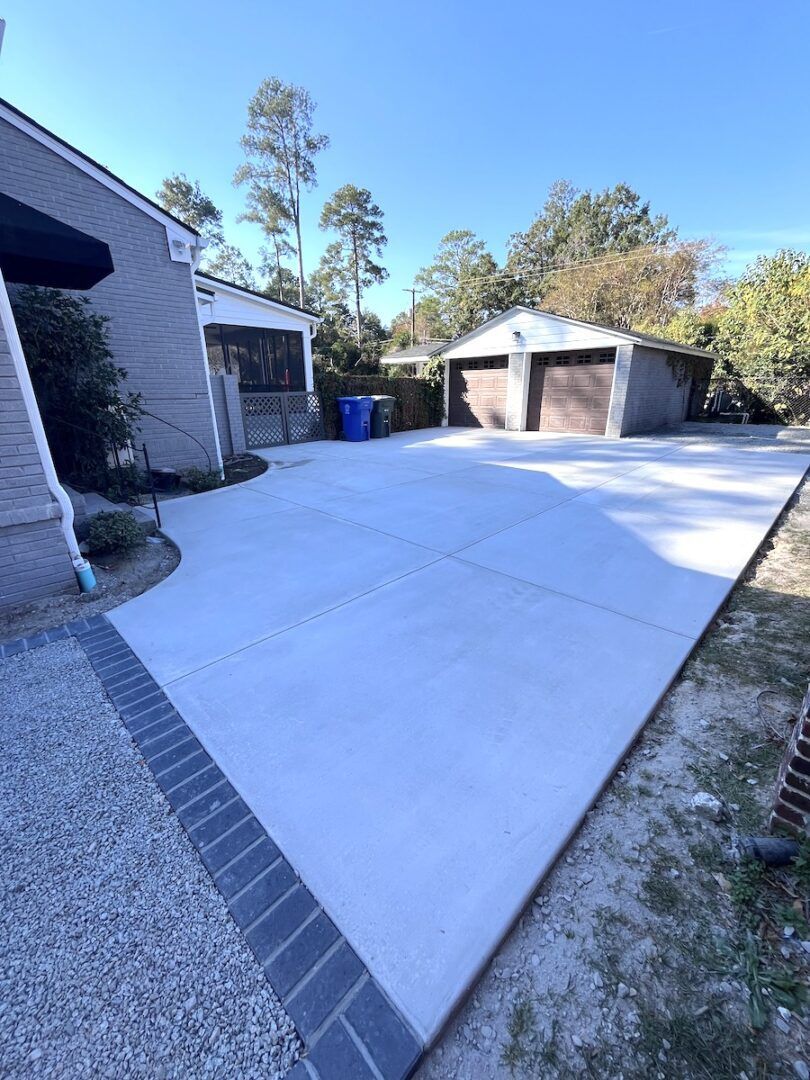 A wide, freshly poured concrete driveway leading to a two-car garage, with a dark brick border and a house to the left.