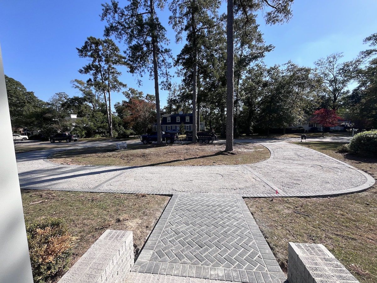 A herringbone-patterned brick walkway leads from a foreground stone structure into a gravel driveway near tall trees.