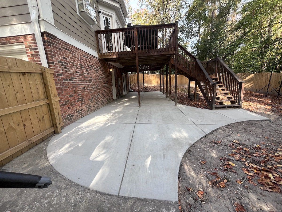 A concrete patio sits beneath a wooden deck with stairs attached to the side of a brick house with a wooden fence.