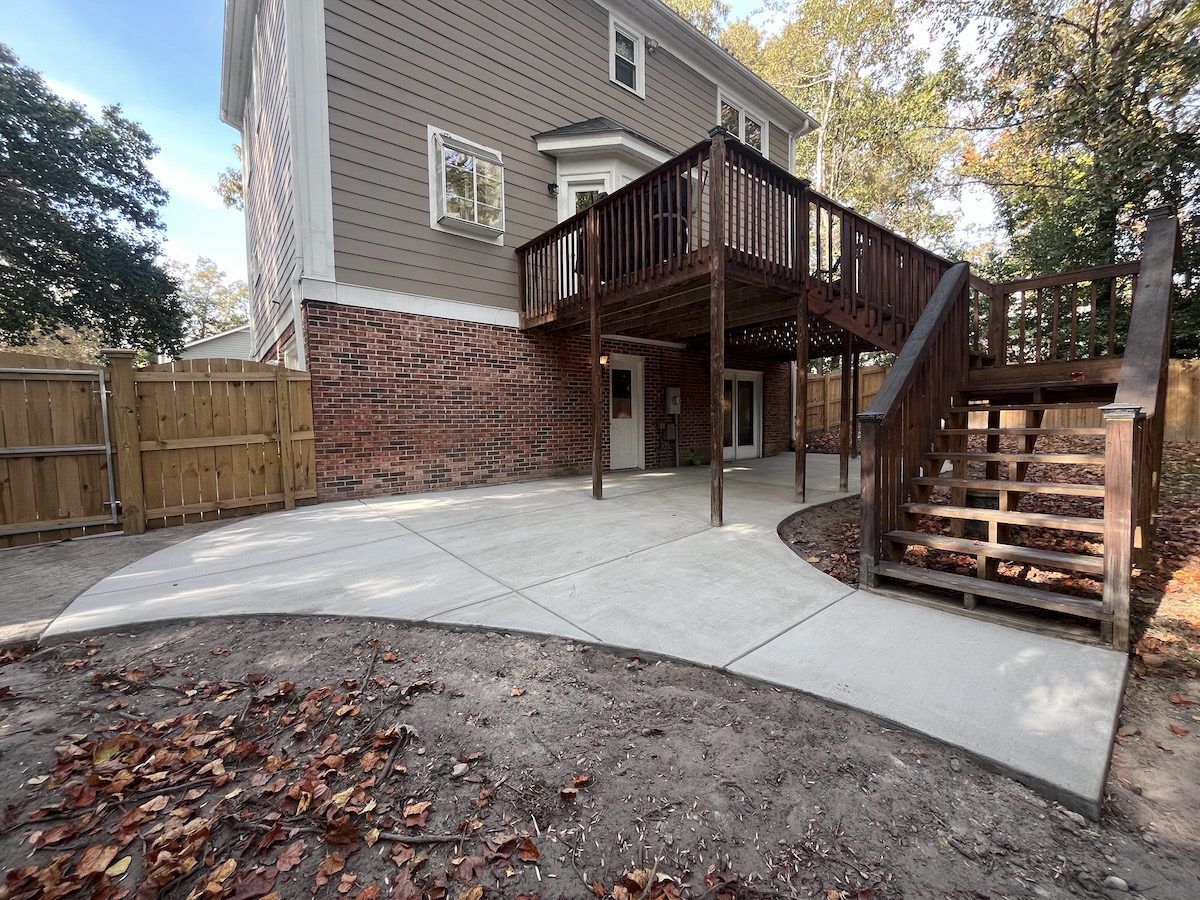A tan house with a brick base, featuring a large wooden deck and a new curved concrete patio in the backyard.