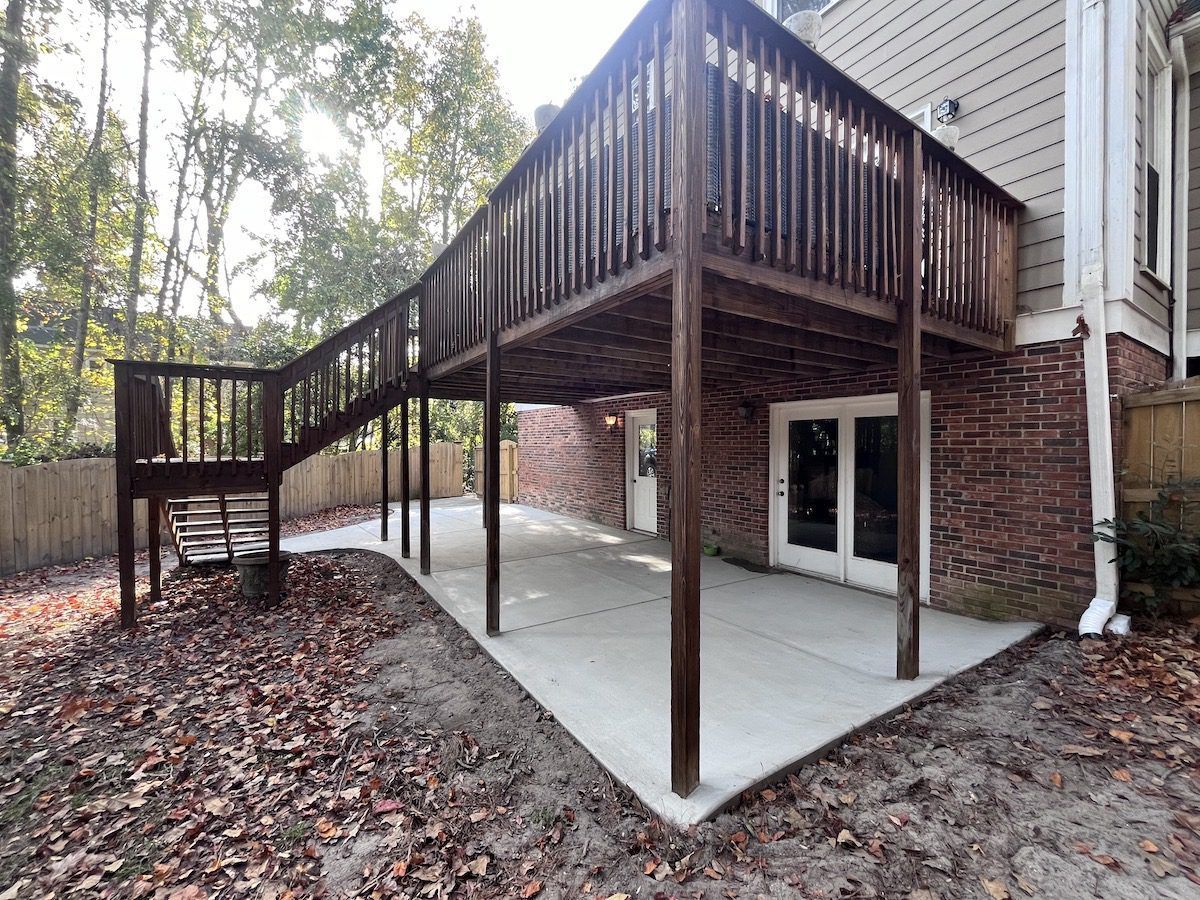 A wood deck with stairs overlooks a concrete patio area behind a brick house surrounded by trees and fallen leaves.