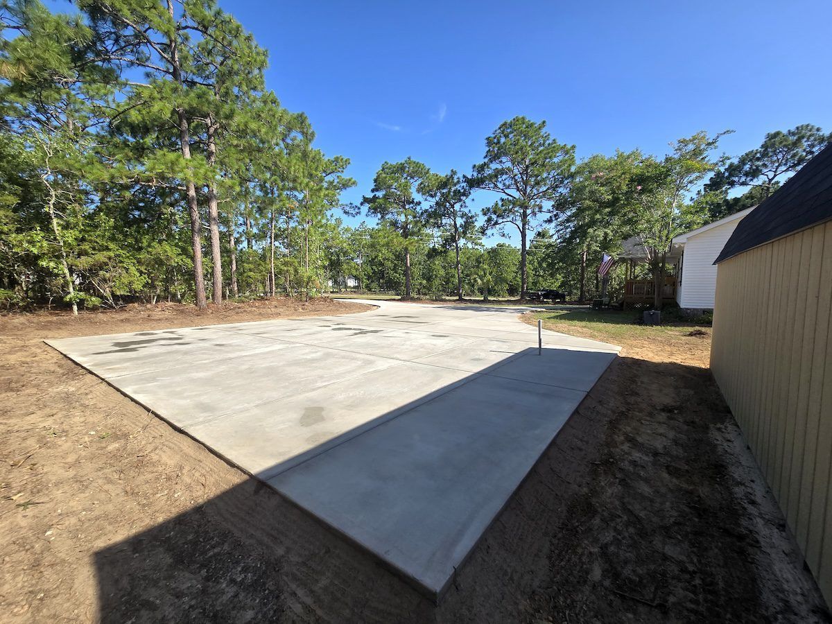 A newly poured concrete slab sits in a wooded backyard next to a light-colored storage shed.