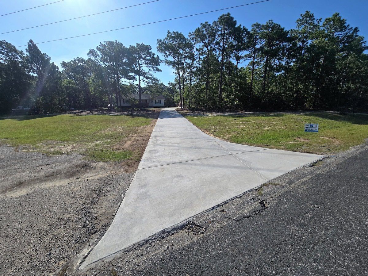 A concrete driveway leading from an asphalt road toward a house nestled among trees under a clear blue sky.