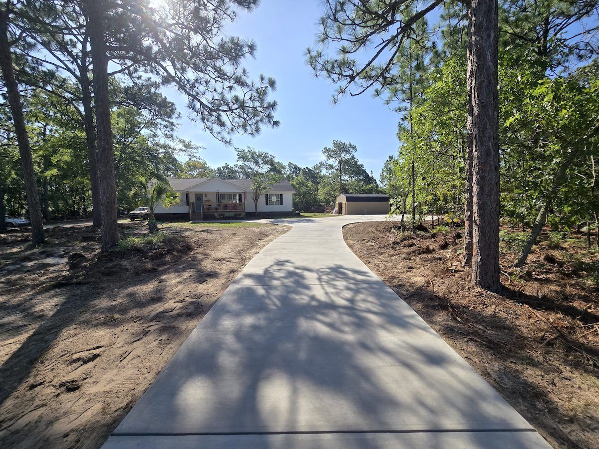 A straight concrete driveway leads through a wooded area toward a single-story house in a sunny, rural setting.