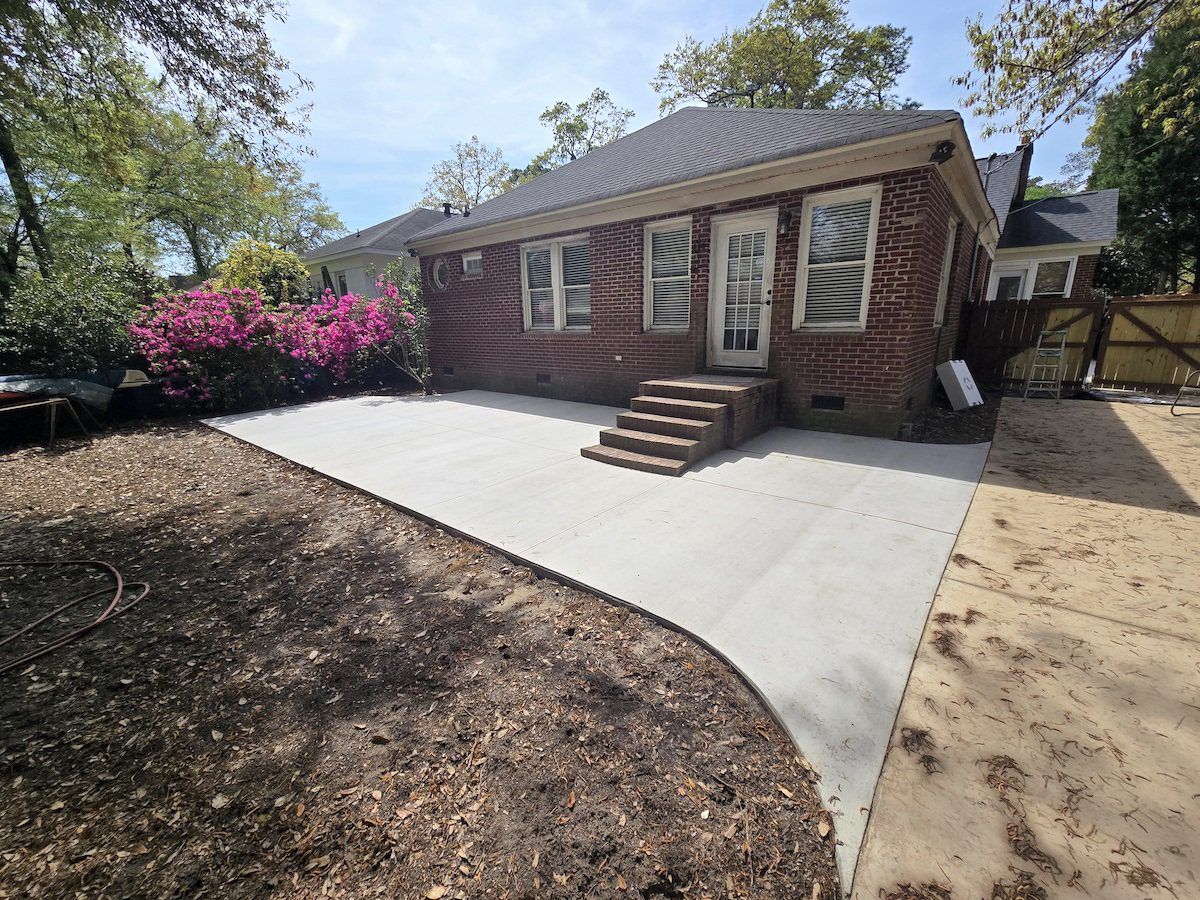 A new rectangular concrete patio at the back of a brick house with stairs leading to a doorway and pink flowering bushes.