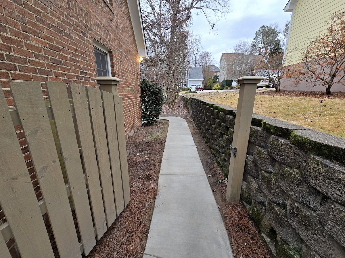 A narrow sidewalk leads between a brick house wall with a wooden fence and a stone retaining wall in a suburban yard.