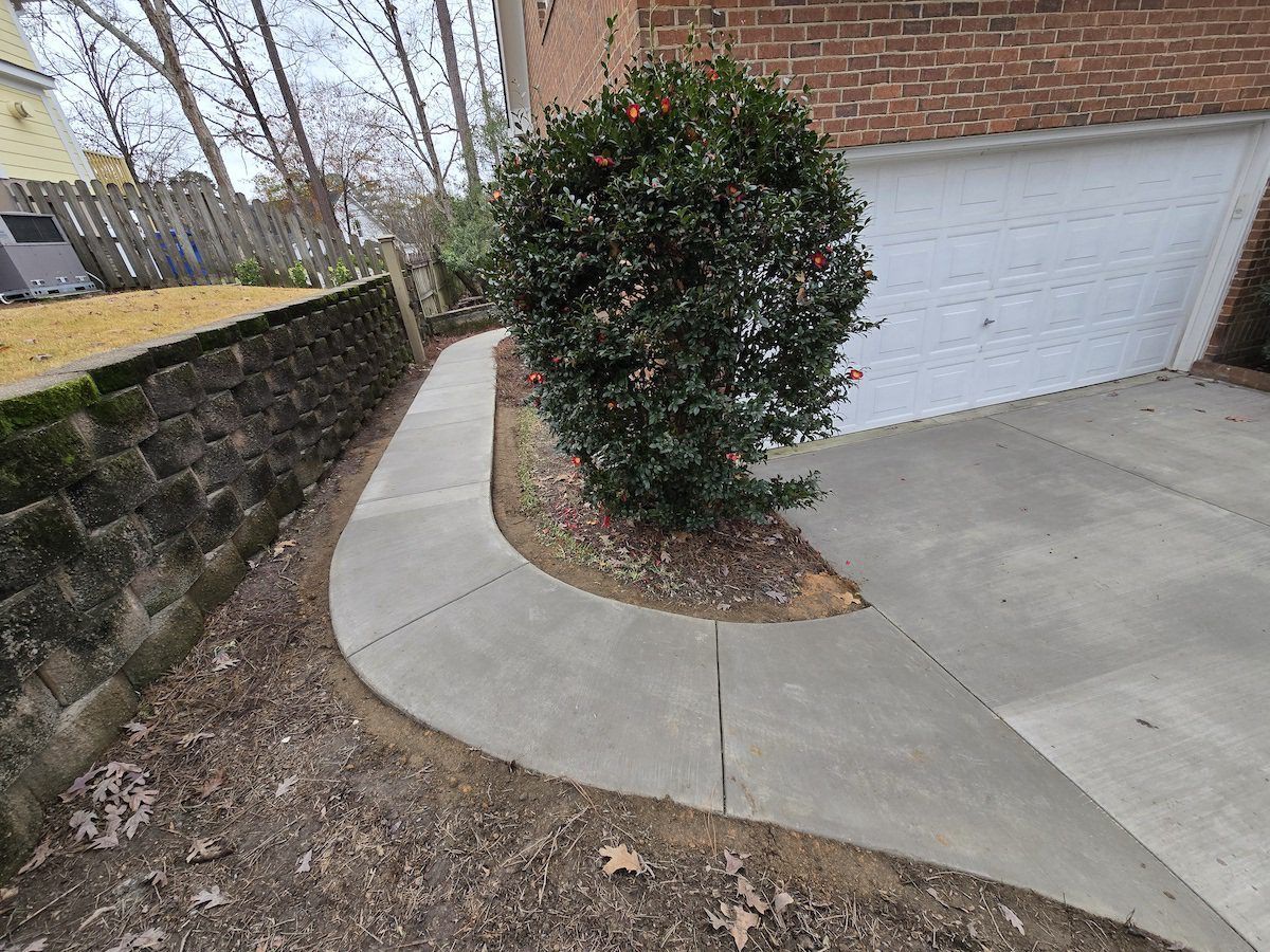 A curved concrete walkway leads along a stone retaining wall past a large bush next to a white garage door.