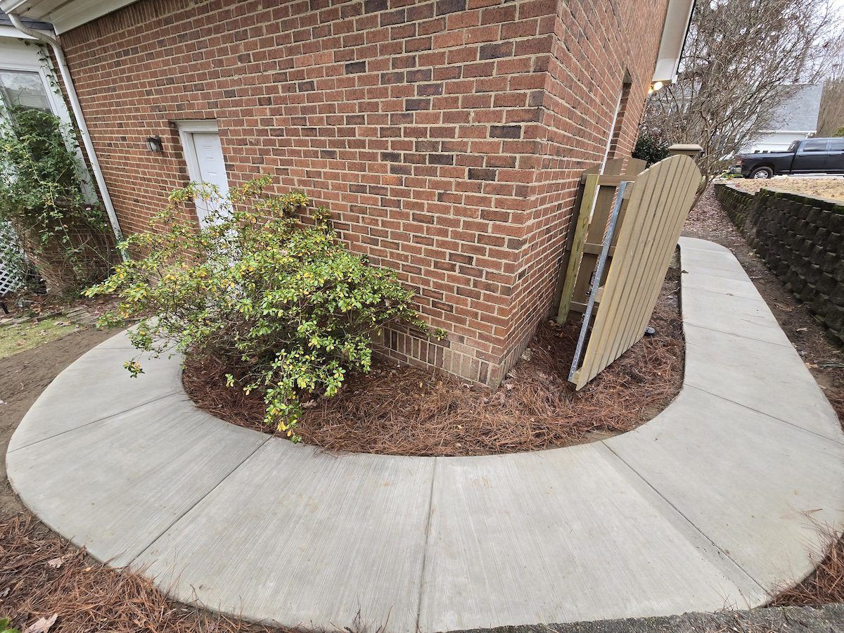 A curved concrete walkway wraps around the corner of a red brick house with a small shrub in the mulched garden bed.