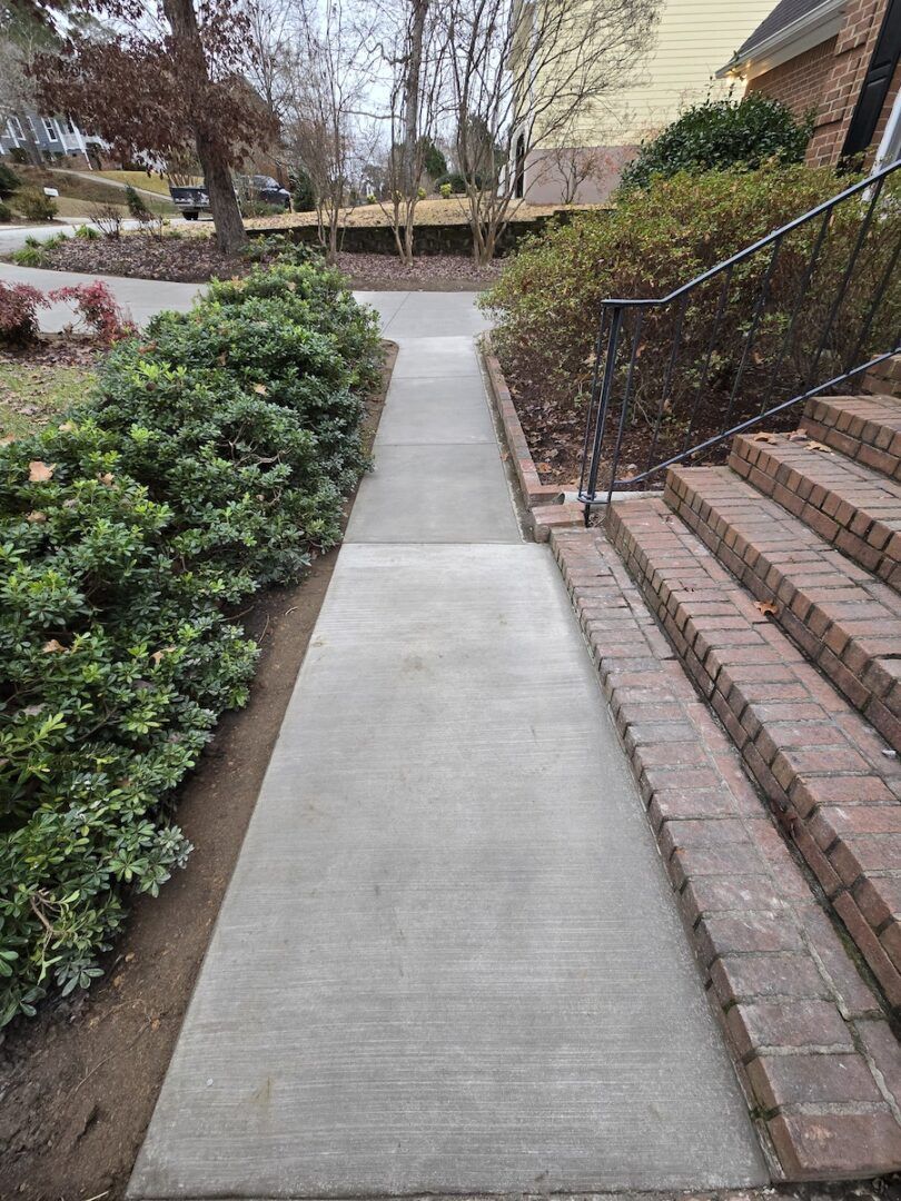 A concrete walkway leads toward a set of brick stairs beside a house, flanked by green bushes on the left.