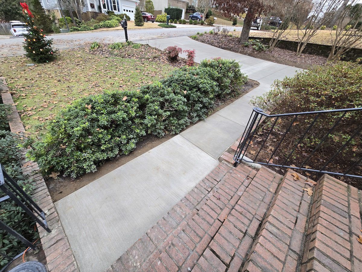 A view from brick steps looking down at a concrete sidewalk, a green hedge, and a suburban street in the background.