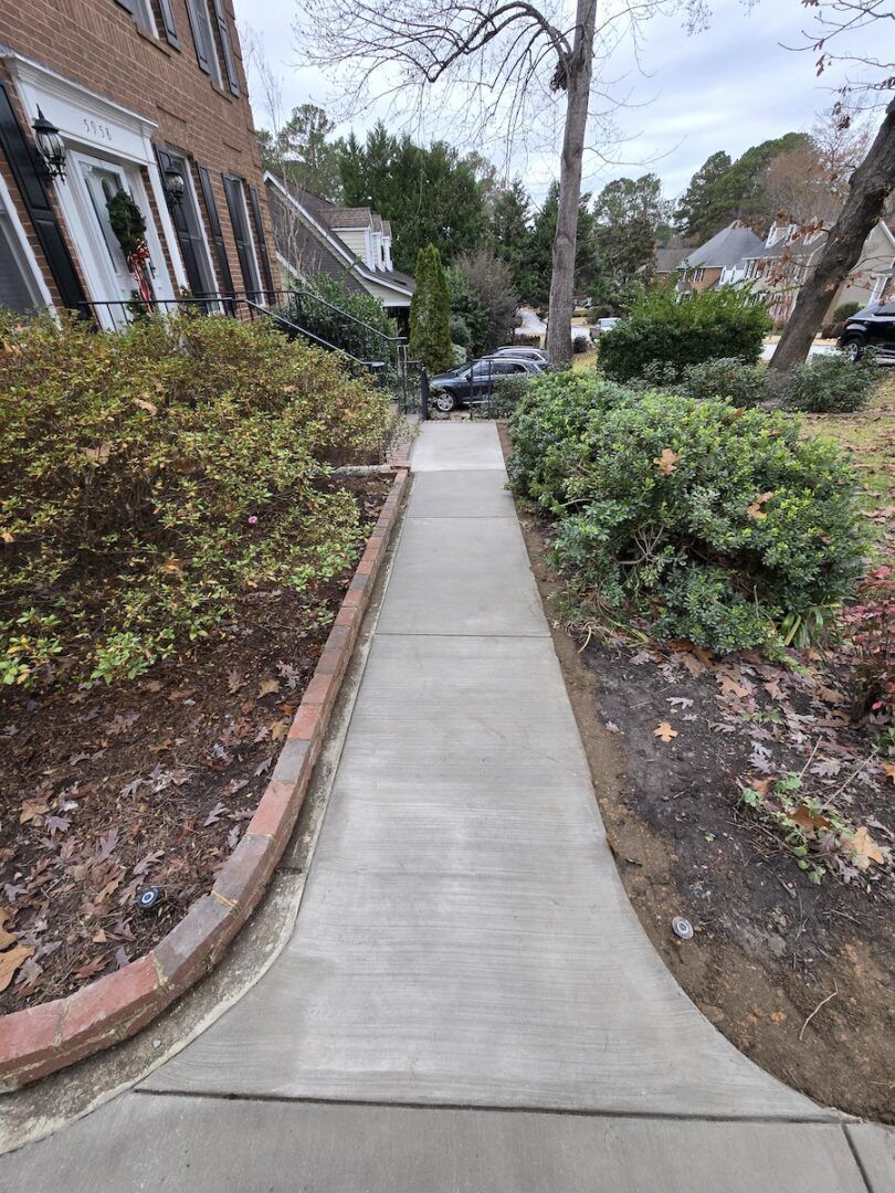 A concrete walkway leads from the bottom center toward a brick house with bushes and a tree in the background.
