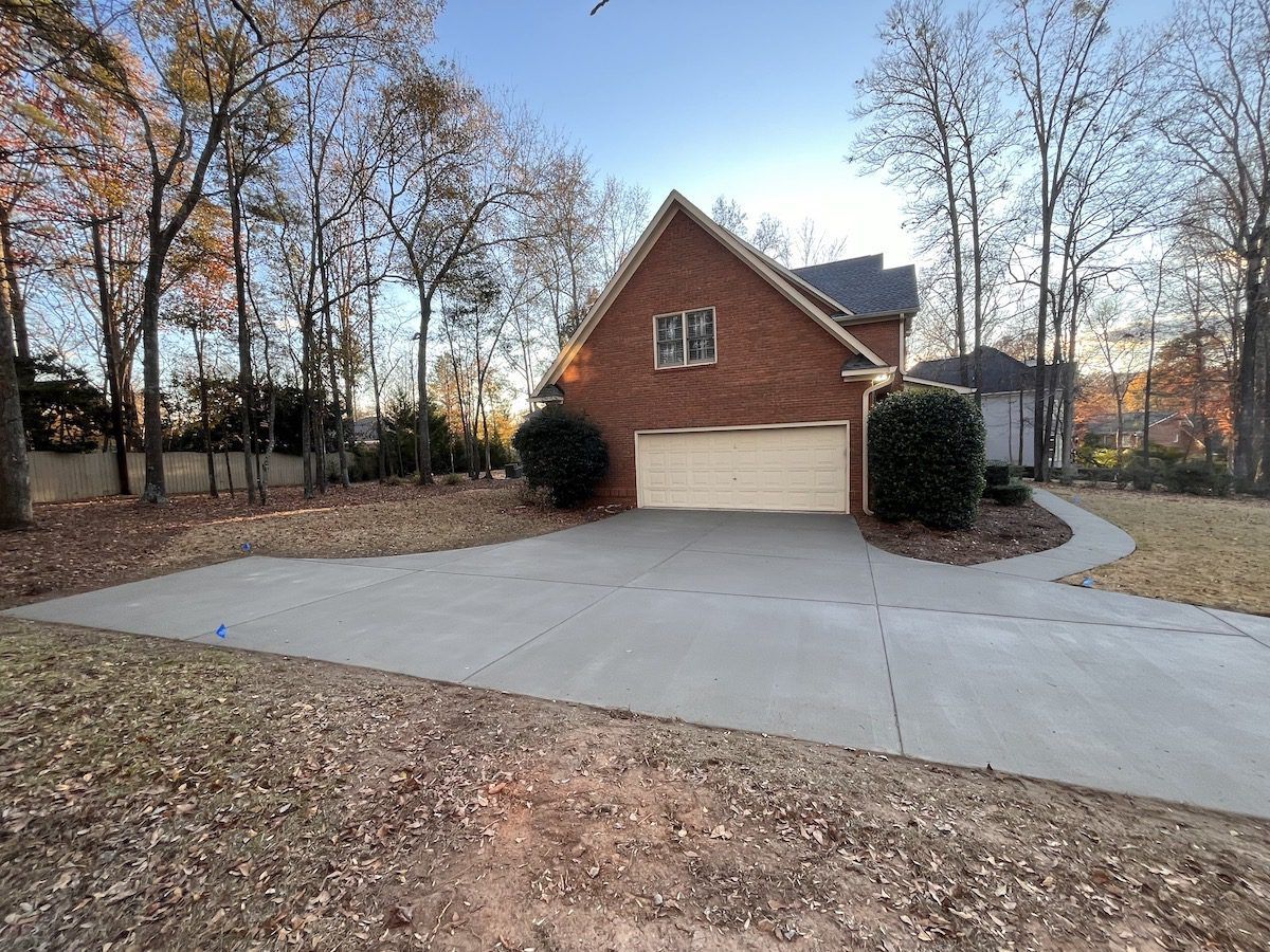 A two-story brick house with a two-car garage at the end of a wide concrete driveway surrounded by trees in autumn.
