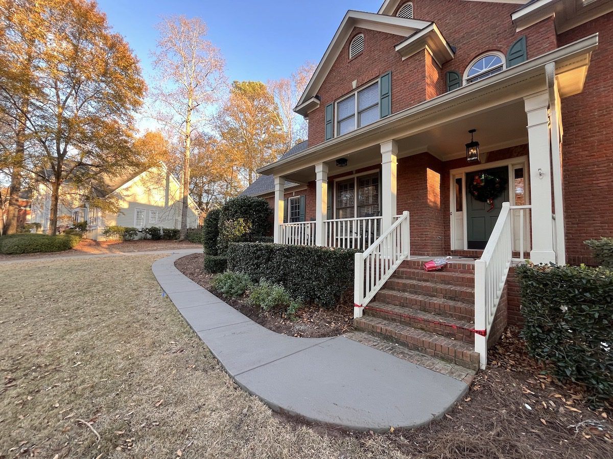 A brick home with a white columned front porch, brick steps, and a concrete walkway leading through a yard in autumn.