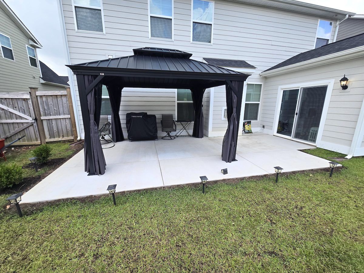 A dark-roofed gazebo sits on a light concrete patio against a multi-story house with a backyard lawn.