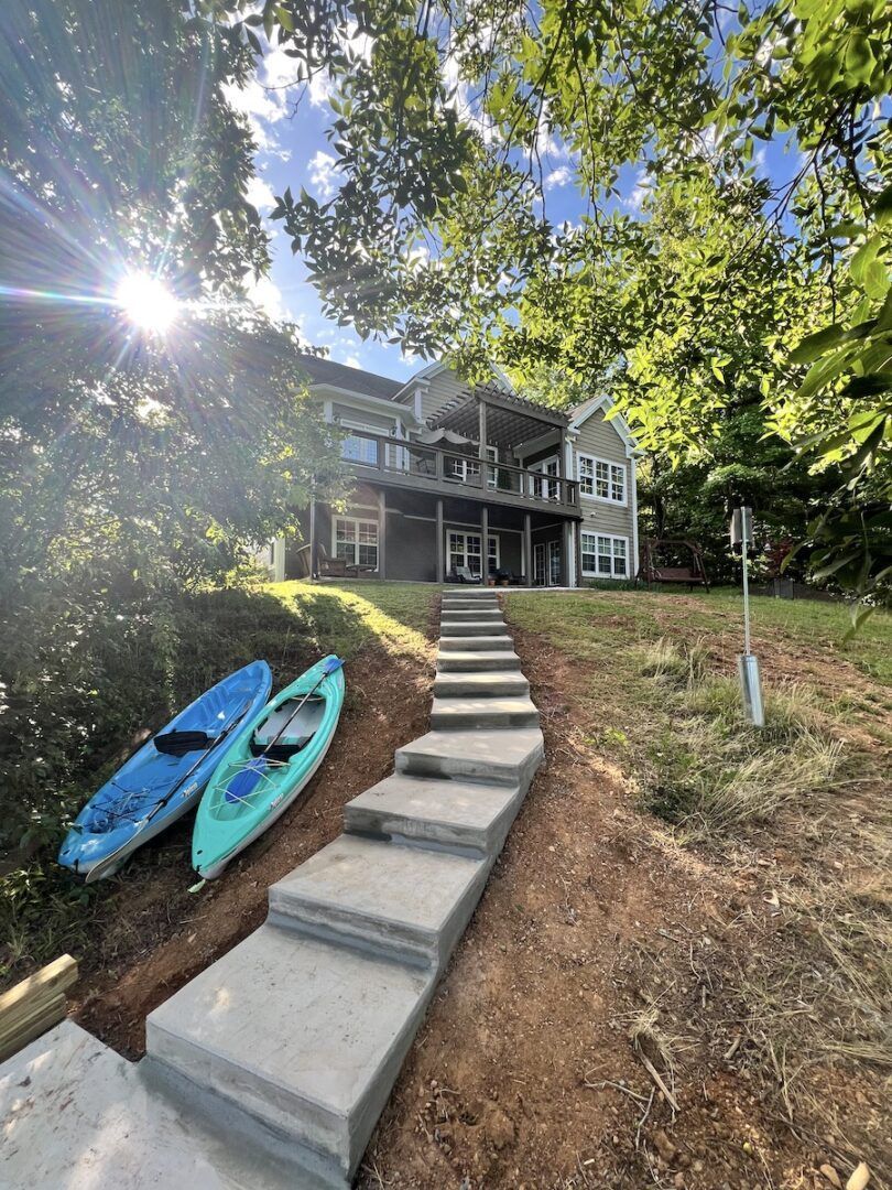 Concrete steps lead uphill toward a two-story house, with two kayaks resting on the grassy slope to the left.