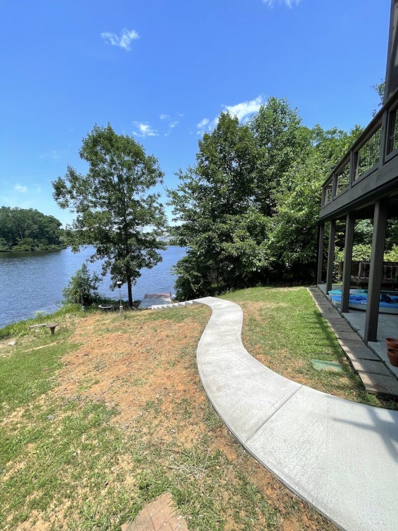 A concrete walkway curves through a grassy yard leading toward a riverbank beside a house deck under a clear blue sky.