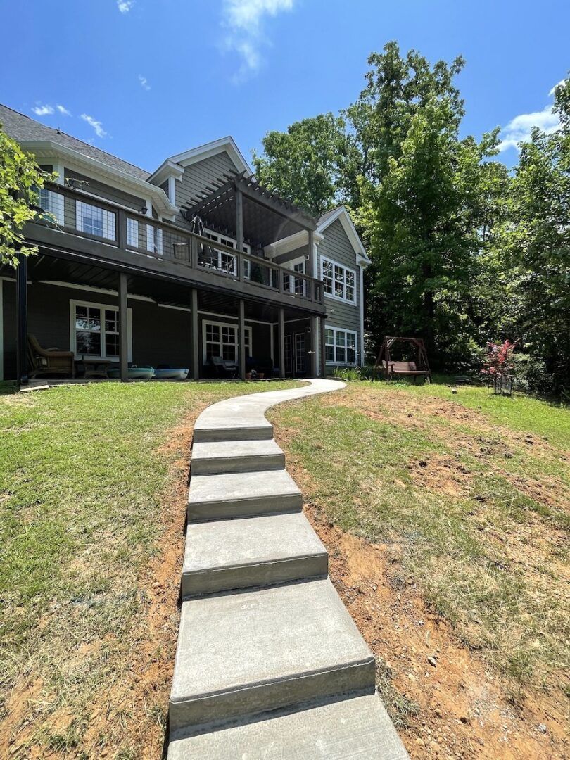 A concrete stairway leads up a grassy hill toward the back of a multi-story house with a wooden deck and forested yard.