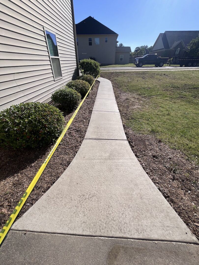 A concrete sidewalk runs along the side of a beige house, lined with green bushes and a yellow caution tape boundary.