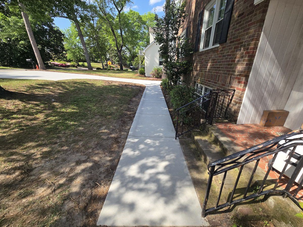 A new, light-gray concrete sidewalk stretches toward a road, running parallel to a brick building's entrance.