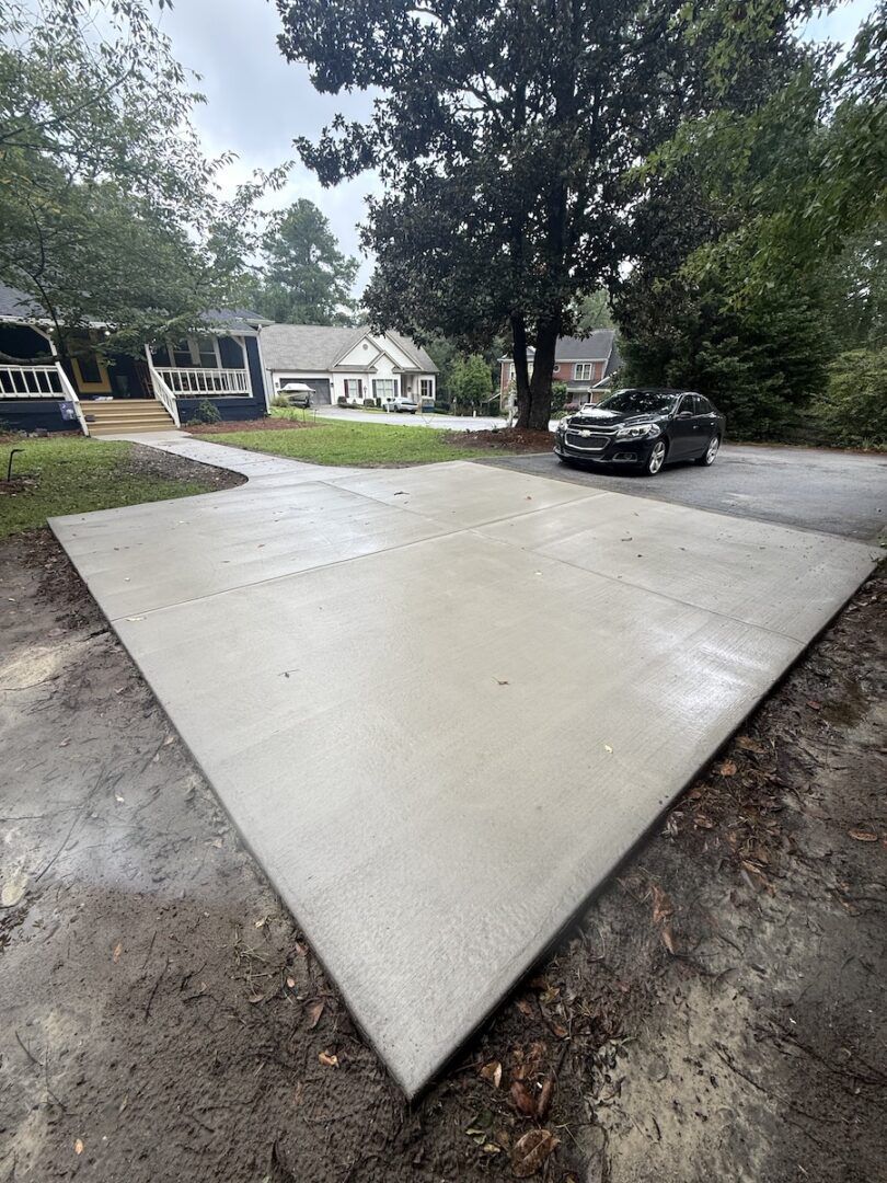 A new square concrete slab sits in a residential yard, connecting to a sidewalk leading to a house with a dark car nearby.