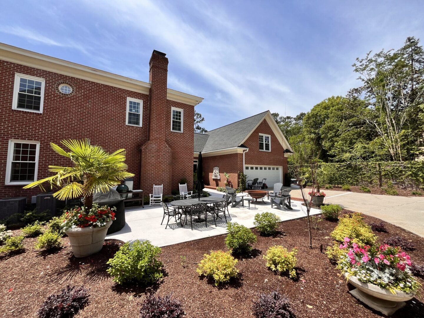 A red brick house with a white patio, outdoor dining set, fire pit, and landscaped garden beds on a sunny day.