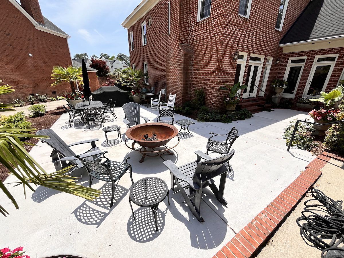A brick house features a spacious light gray patio furnished with metal chairs, small tables, and a central fire pit.