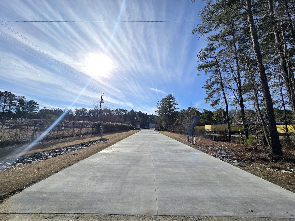 A straight concrete driveway stretches toward a line of trees under a bright, sunny sky with wispy clouds.