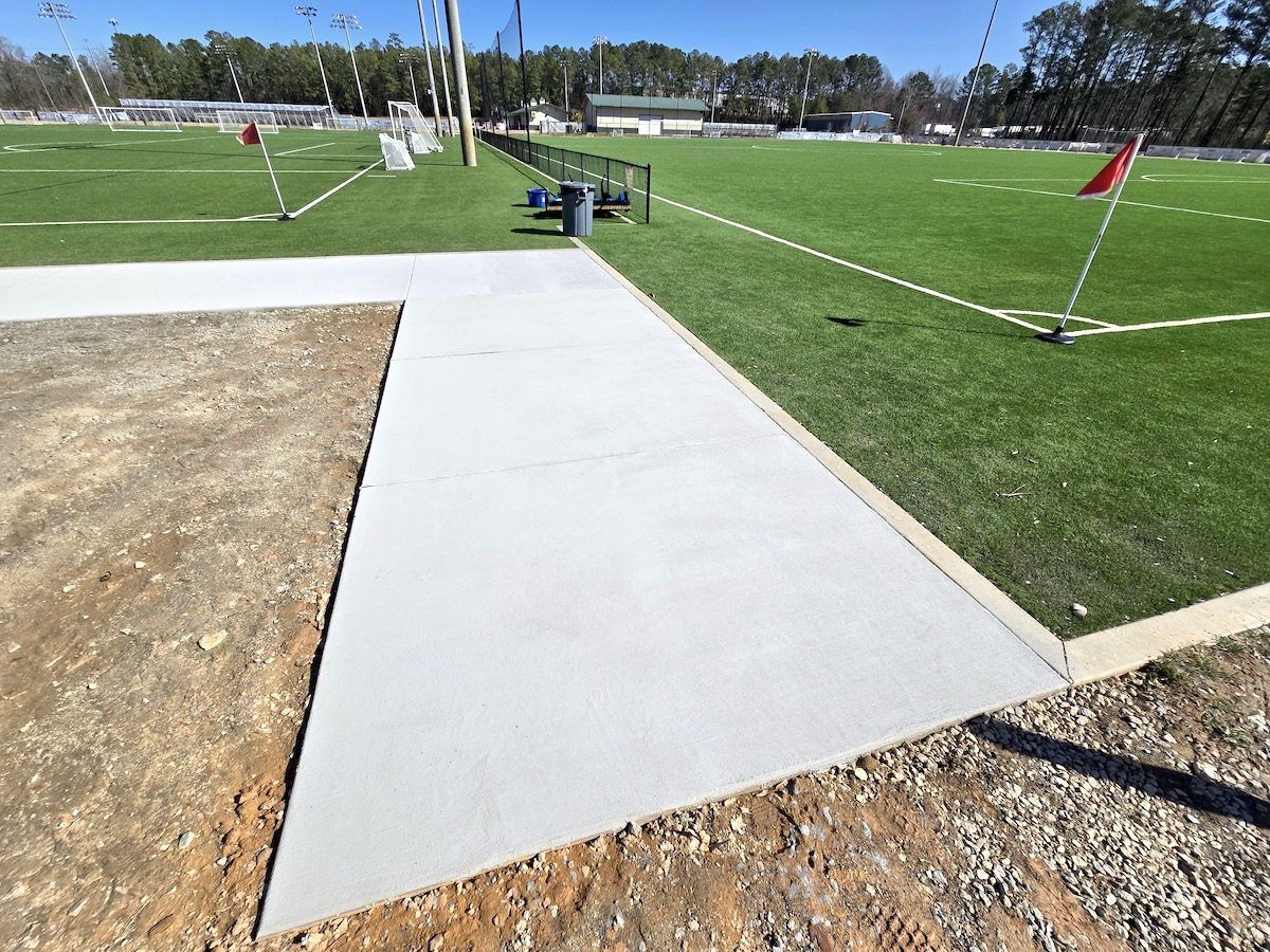A concrete walkway borders a grassy field and a dirt area, with a fence and sports field in the background.