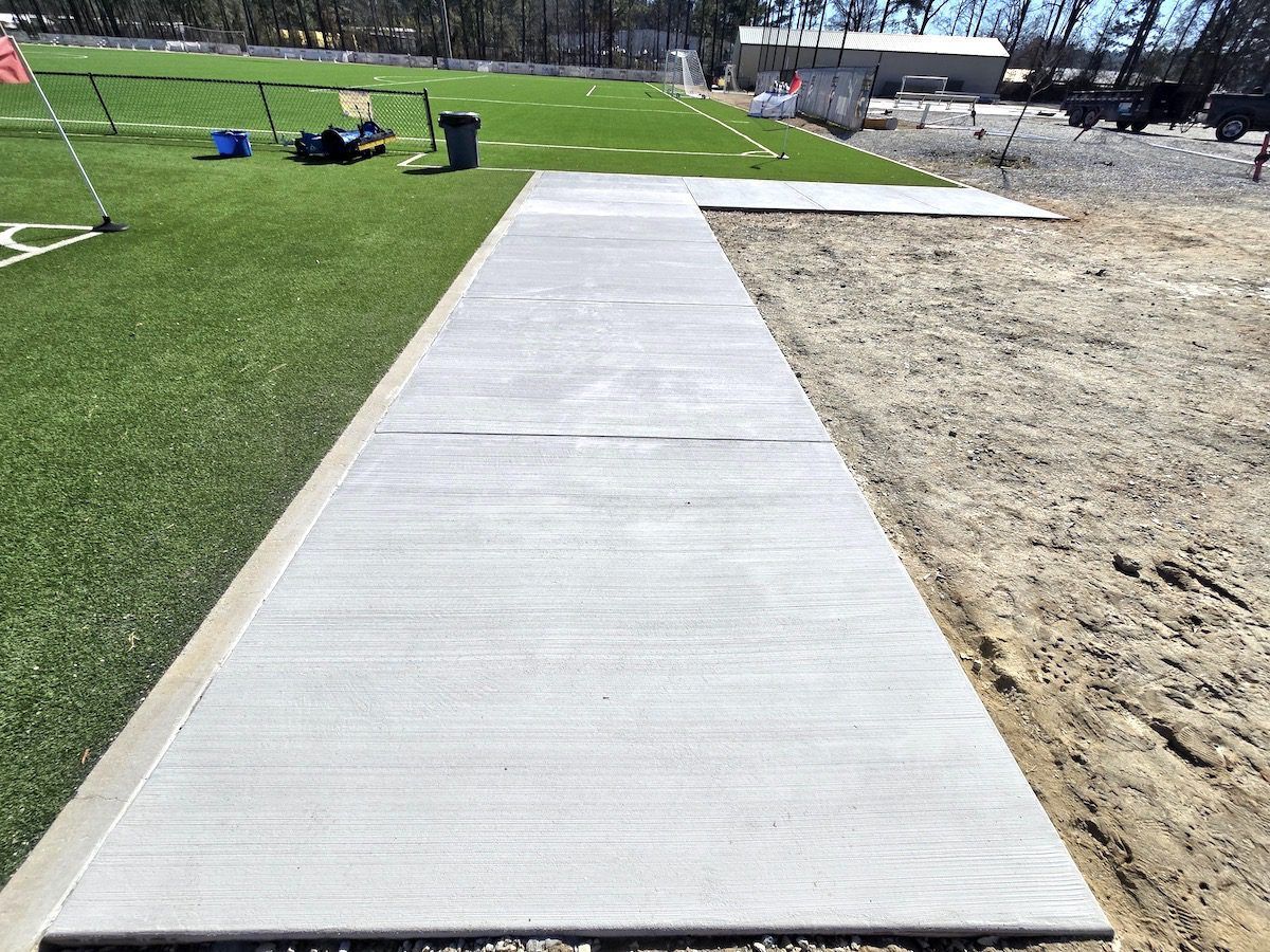 A long, light gray concrete walkway runs alongside a green artificial turf field and an area of packed dirt.