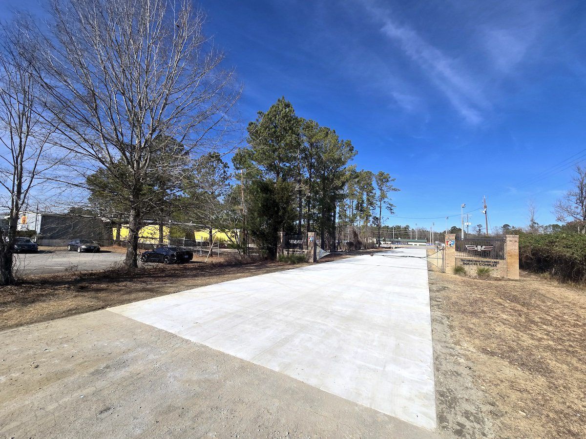 A newly paved white concrete path leads toward a brick entryway gate under a bright blue sky with trees and a parking lot.