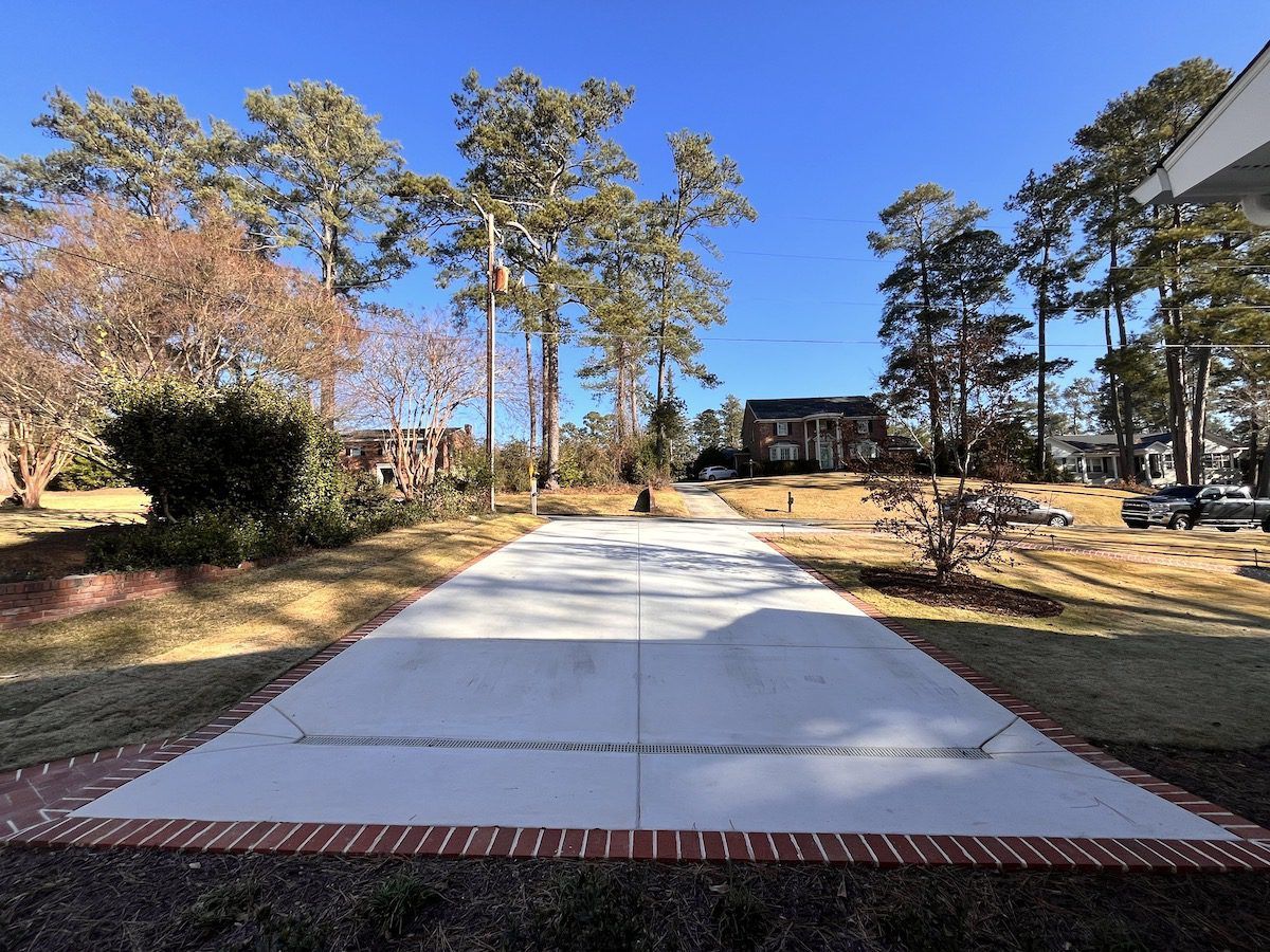 A concrete driveway with a brick border leads toward a house surrounded by tall pine trees on a sunny day.