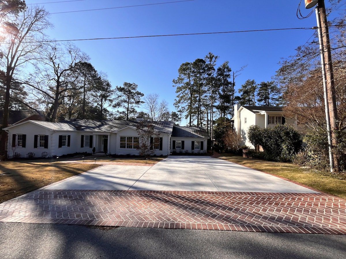 White ranch-style house with a large concrete and brick driveway, surrounded by trees under a clear blue sky.