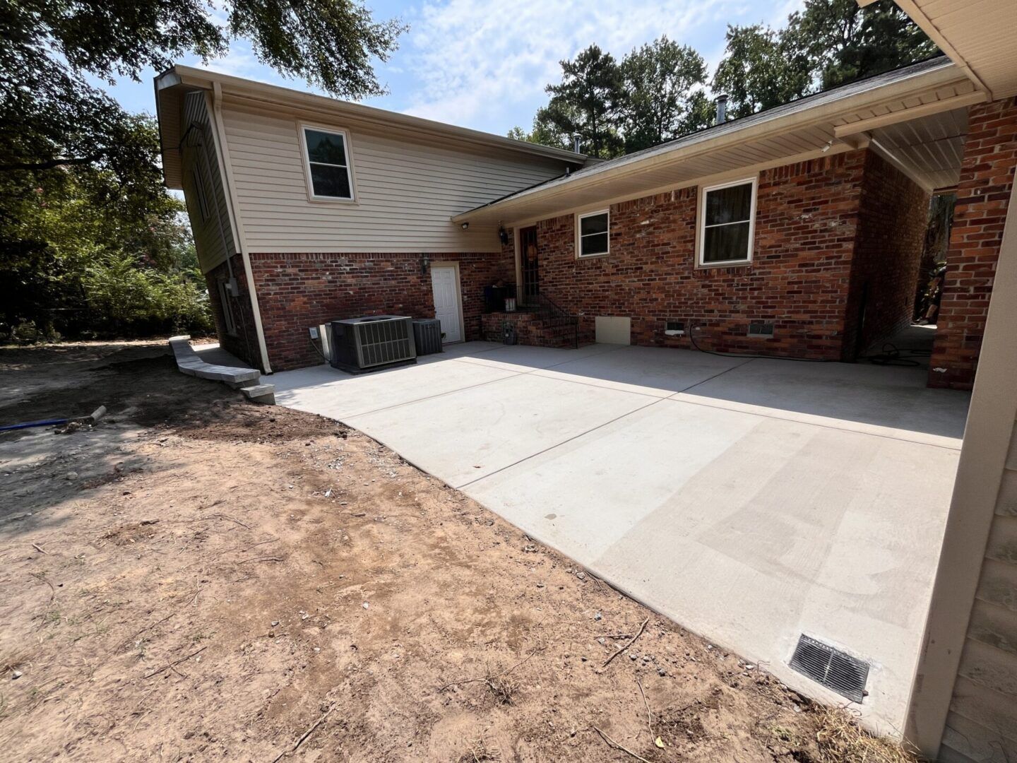 A newly poured concrete patio adjacent to a brick and siding house with an outdoor HVAC unit and an unlandscaped yard.