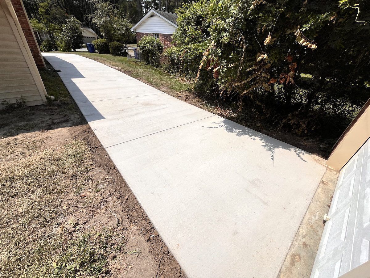 A concrete driveway extends from the edge of a house toward the street, flanked by grass and a shrubbery line.