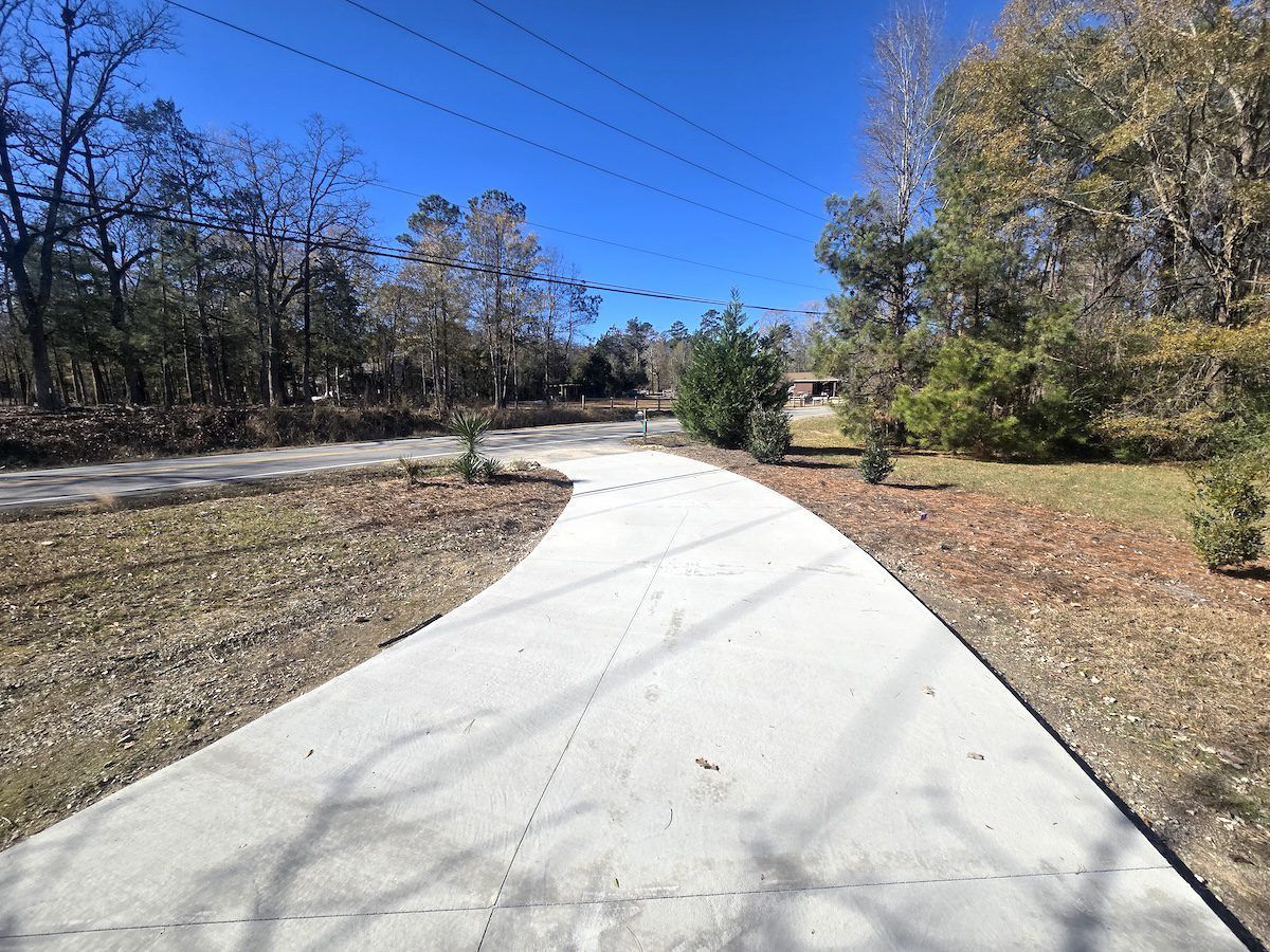A concrete driveway curves through a residential yard with bare trees, dry leaves, and a clear blue sky.