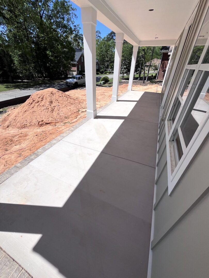 A view along a concrete residential porch with white columns, beside a house with grey siding and a large mound of dirt.