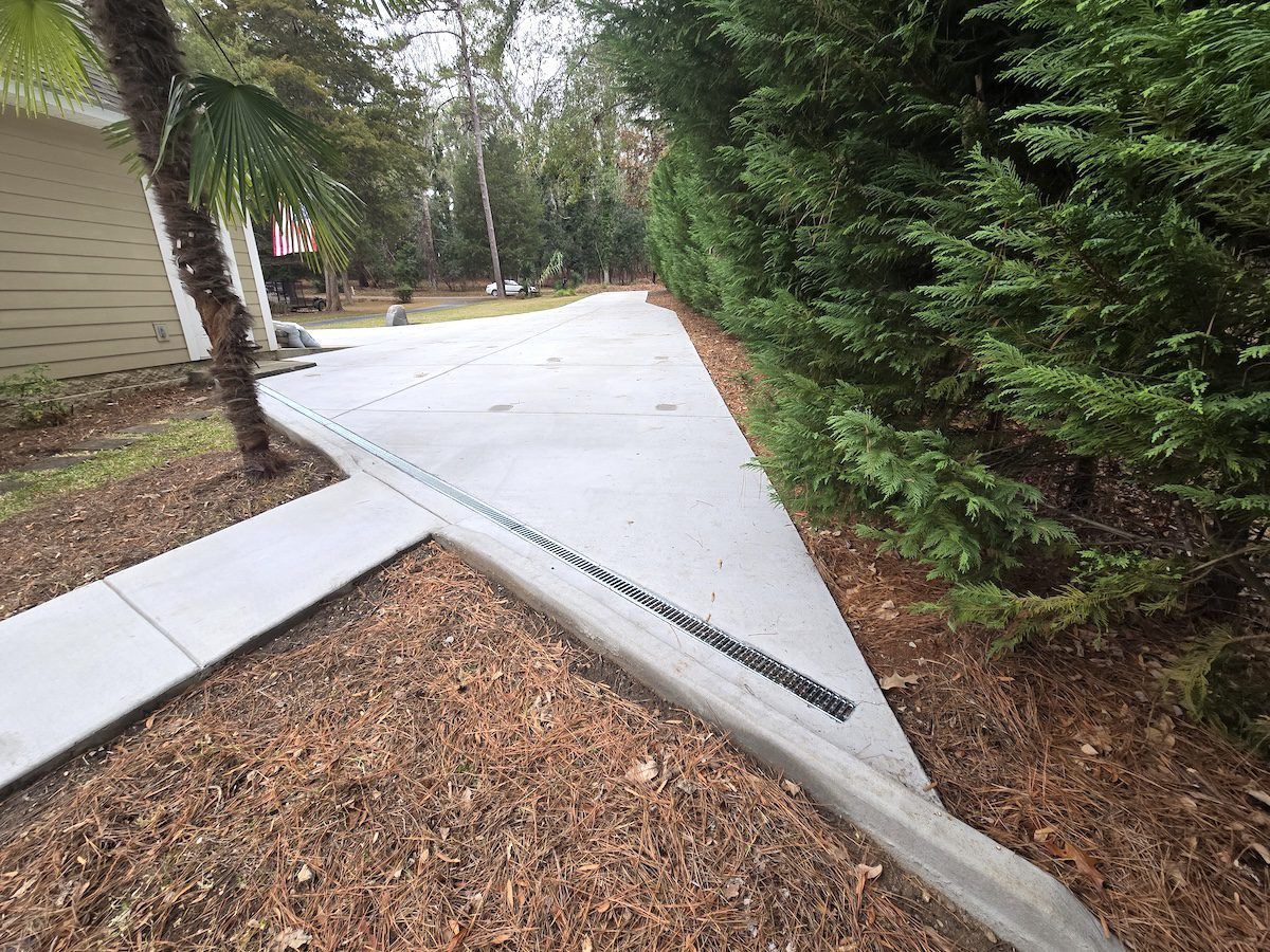 A concrete driveway featuring a built-in trench drain across its surface, surrounded by pine straw mulch and dense greenery.
