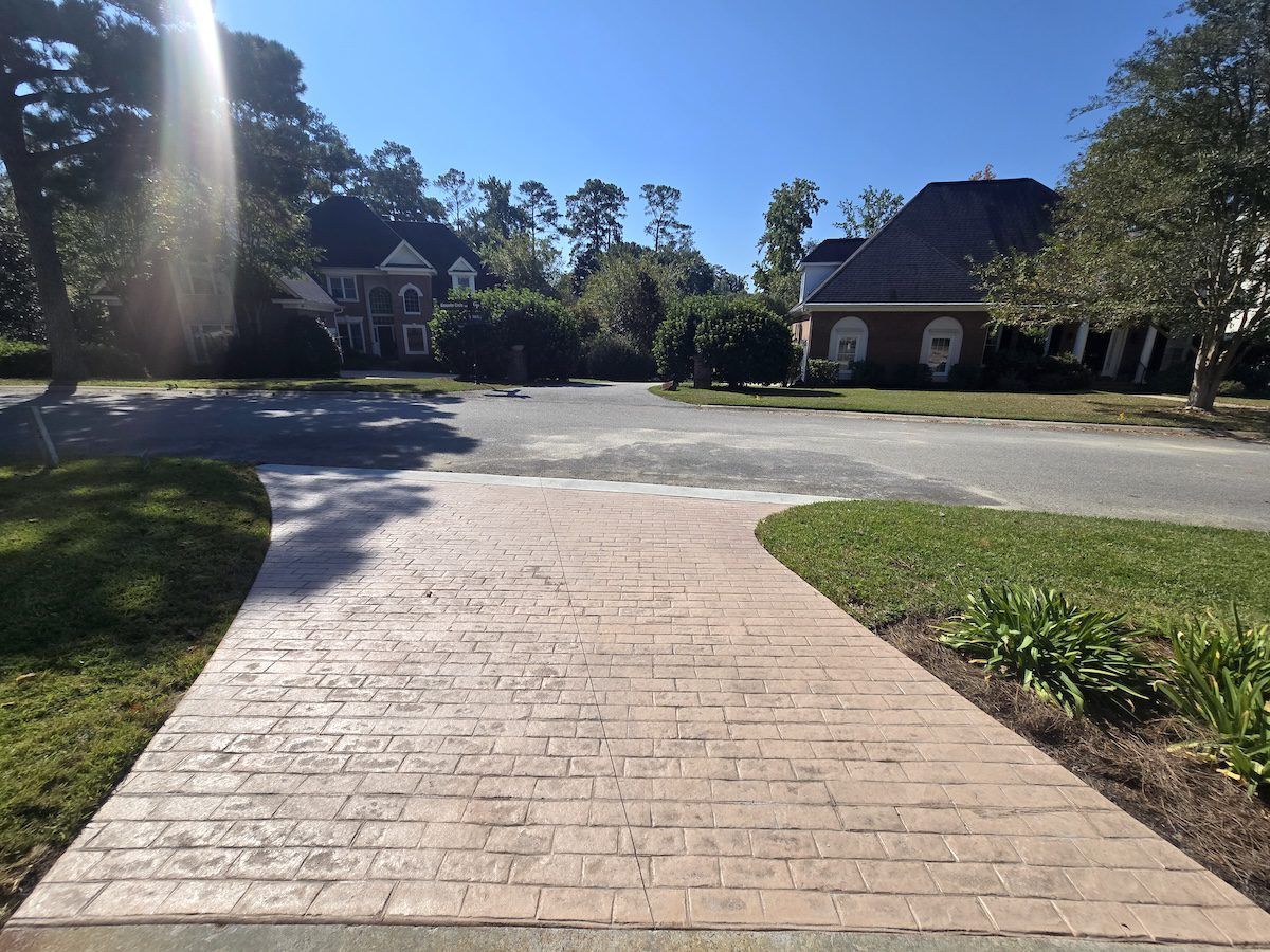 A tan, textured concrete driveway with a brick-like pattern leads toward a cul-de-sac with homes surrounded by trees.