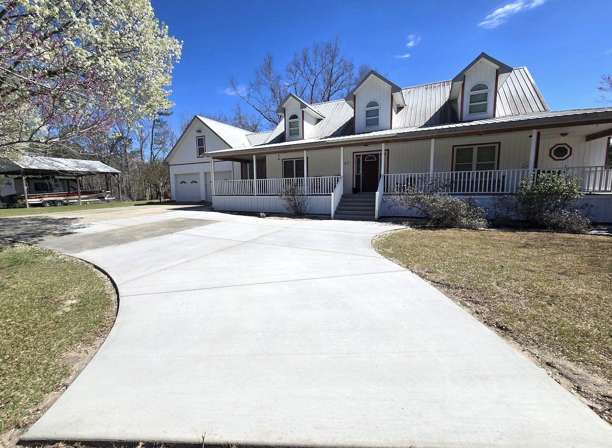 A large white farmhouse with a metal roof and a wide concrete driveway under a bright blue sky.