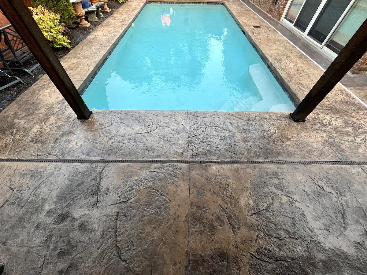 A bright blue rectangular swimming pool surrounded by a weathered, textured concrete patio under a wooden pergola.