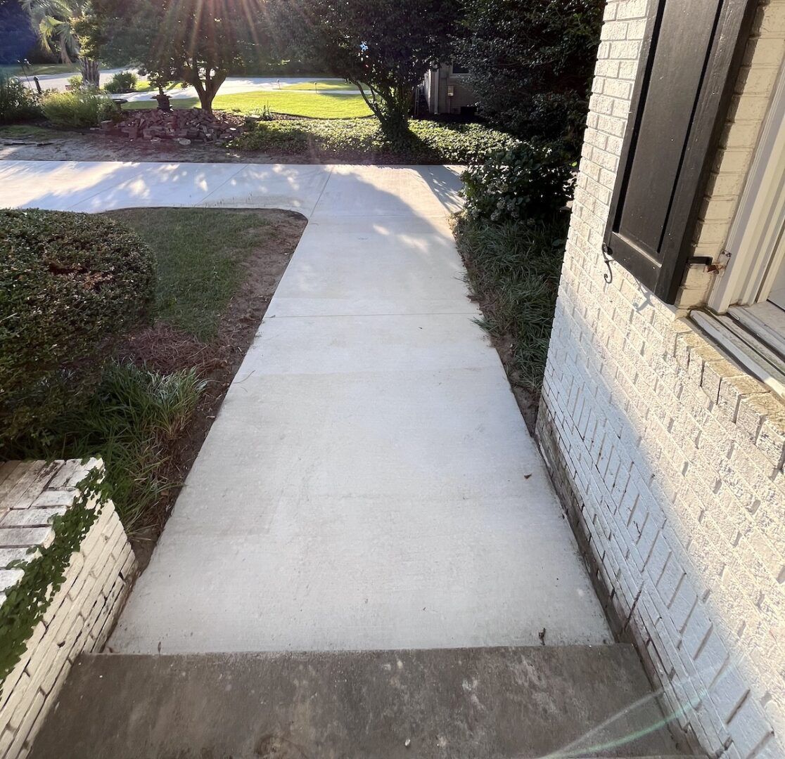 A concrete sidewalk extends from a doorway, flanked by a white brick wall and green landscaping on a sunny day.