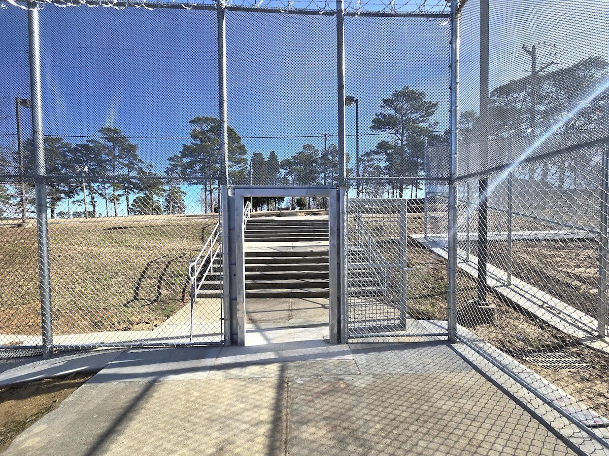 A paved path leads through a metal gate and up a set of concrete stairs enclosed by tall chain-link fencing.