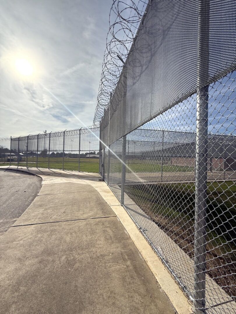 A chain-link security fence topped with coiled razor wire runs along a concrete path under a bright, sunny sky.