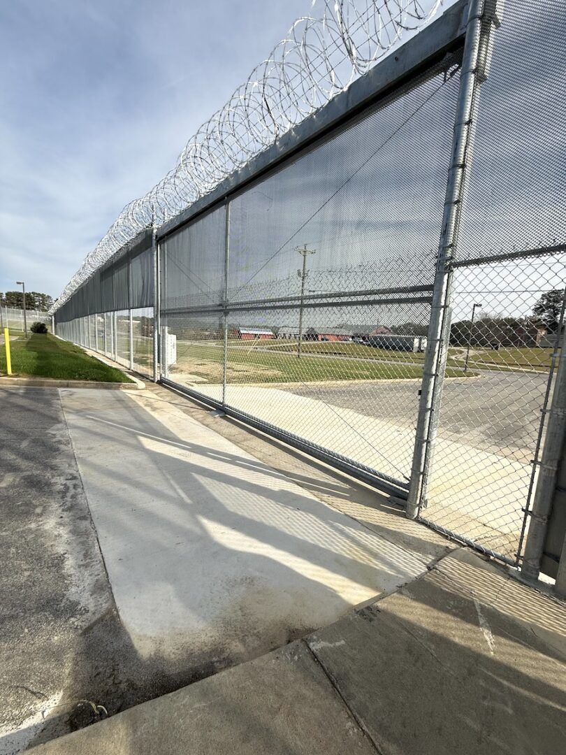 A tall chain-link security fence topped with razor wire runs alongside a paved path on a bright, sunny day.
