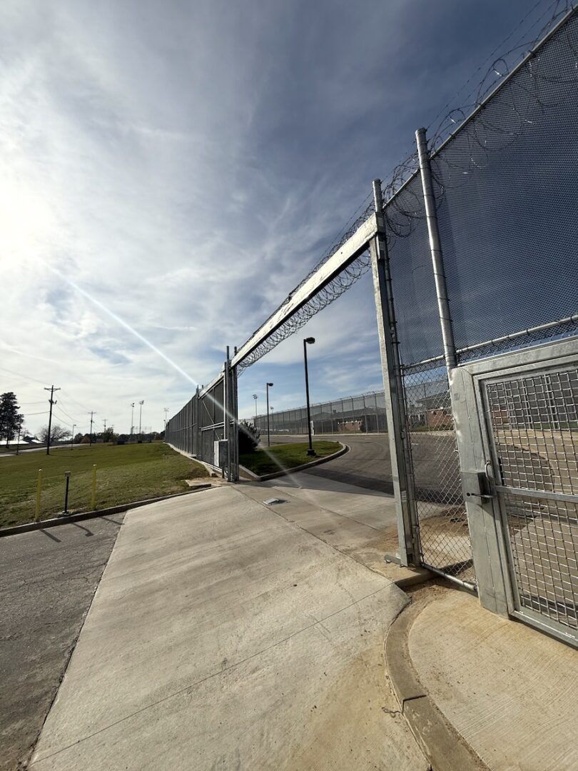 A low-angle view of a tall metal chain-link fence topped with razor wire, stretching across a sunny outdoor perimeter.